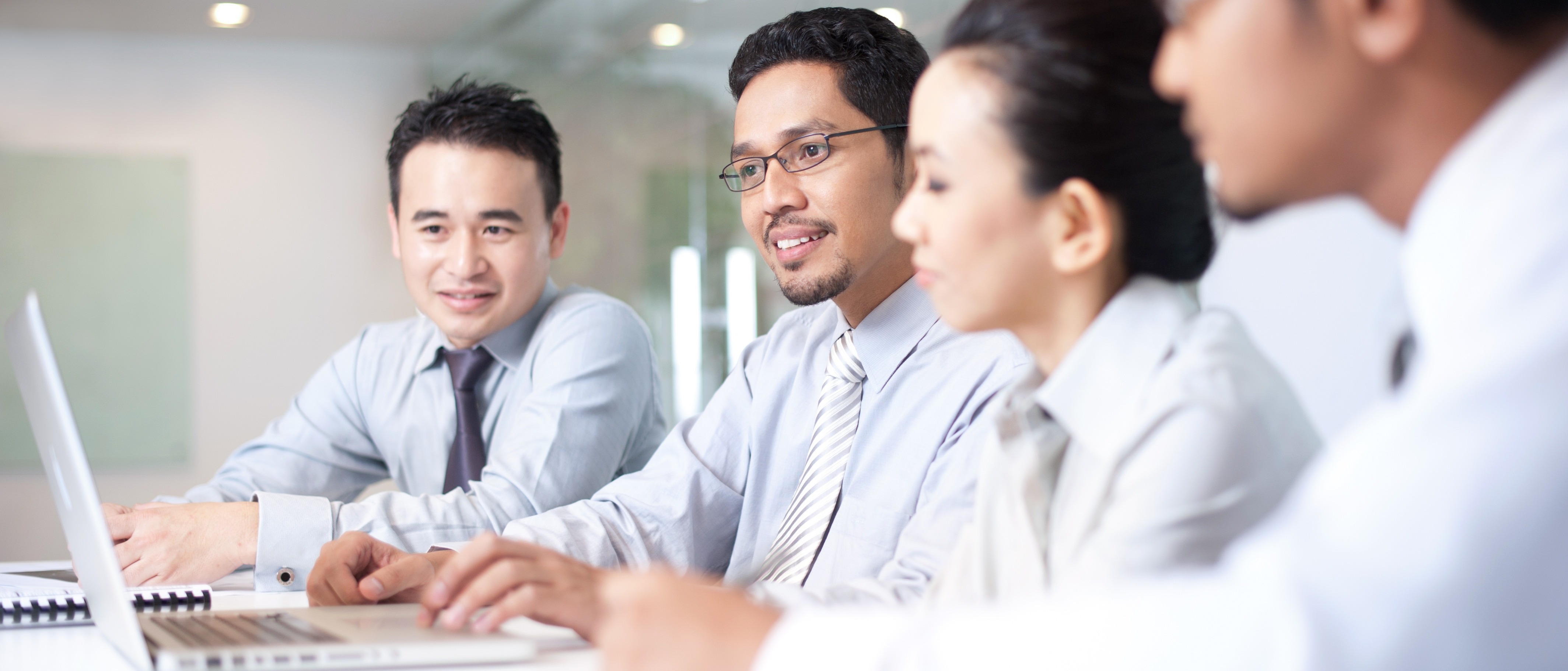 Four people in business attire sit at a conference table, focused on a laptop.